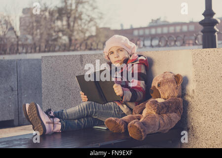 Kleines Mädchen sitzt auf einer Bank und liest das Buch um ein Spielzeug Bär. Verarbeitung: Muskelaufbau in warmen Farben Stockfoto