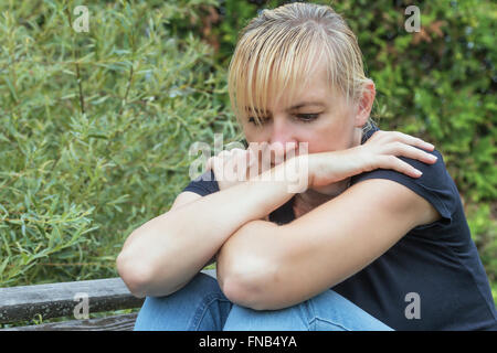 Frustriert blonde Frau ist im schwarzen Hemd auf der Holzbank im Freien sitzen. Ihre Hände ruhen auf den Knien. Stockfoto