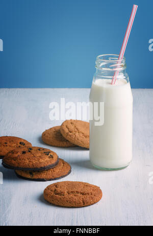 Schokoladenkekse mit einer Milchflasche mit einem Strohhalm auf einem weißen Holztisch und blauem Hintergrund. Stockfoto