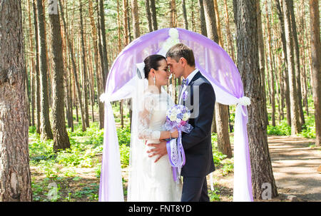 Wunderschöne stilvolle reich glücklich Braut und Bräutigam stehen bei einer Trauung im Garten in der Nähe von lila Bogen mit Blumen Stockfoto