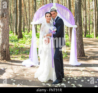 Wunderschöne stilvolle reich glücklich Braut und Bräutigam stehen bei einer Trauung im Garten in der Nähe von lila Bogen mit Blumen Stockfoto