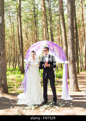 Wunderschöne stilvolle reich glücklich Braut und Bräutigam stehen bei einer Trauung im Garten in der Nähe von lila Bogen mit Blumen Stockfoto