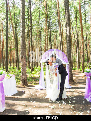 Wunderschöne stilvolle reich glücklich Braut und Bräutigam stehen bei einer Trauung im Garten in der Nähe von lila Bogen mit Blumen Stockfoto