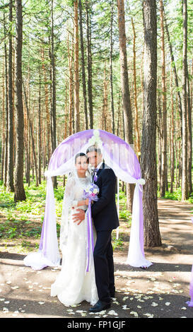 Wunderschöne stilvolle reich glücklich Braut und Bräutigam stehen bei einer Trauung im Garten in der Nähe von lila Bogen mit Blumen Stockfoto