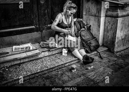 eine Dame, die schon bessere Tage sitzen auf einer Tür Stufen Rollen eine Zigarette, mit ihren Habseligkeiten in einen Rucksack gesehen hat. Stockfoto