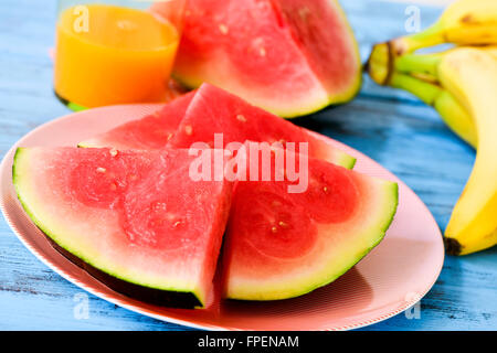 Nahaufnahme einer Platte mit einigen Stücken von erfrischenden Wassermelonen, Bananen und ein Glas Orangensaft auf einen blauen rustikalen hölzernen t Stockfoto