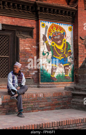 Nepal, Patan Durbar Square.  Mann sitzt neben einem Gemälde Bhairab (Bhairav), eine heftige Manifestation des Gottes Shiva. Stockfoto