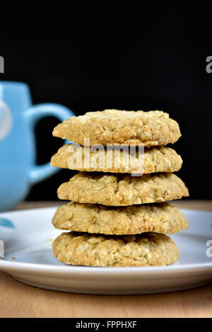 Haufen von hausgemachten Hafer Cookies auf Teller mit Rand des blauen Becher vor schwarzem Hintergrund Stockfoto