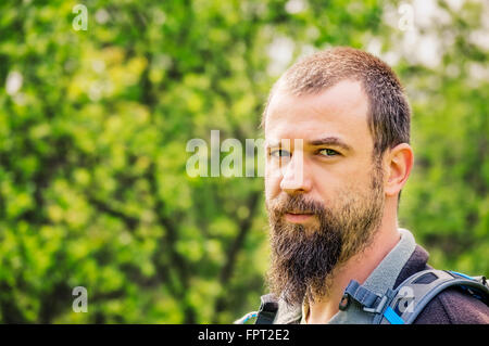 Porträt eines bärtigen jungen Mannes mit Rucksack mit grünen Bäumen im Hintergrund. Stockfoto