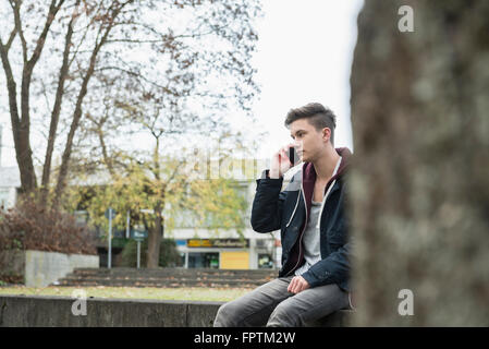 Junger Mann telefonieren mit Handy und sitzen auf Sims, München, Bayern, Deutschland Stockfoto