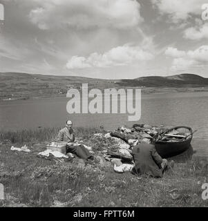 Historisches Bild der 1950er Jahre, Bild des englischen Fotografen J Allan Cash, der ein Picknick an einem See in Irland mit einem ortsansässigen Mann in Begleitung macht. Ein kleines Ruderboot aus Holz ist am Ufer festgemacht. Stockfoto