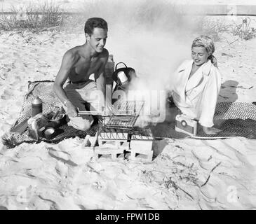 1940ER JAHREN PAAR PICKNICK AM STRAND MANN GRILLEN HOT DOG Stockfoto