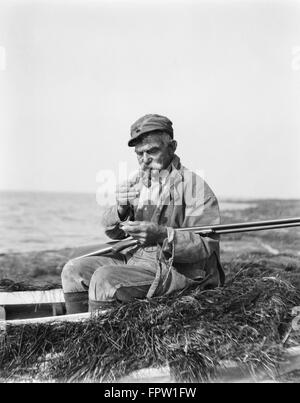 1920ER JAHRE MANN BELEUCHTUNG CORN COB PIPE SITZEN IN BARNEGAT BAY SNEAKBOX BOOT IN SÜMPFEN MIT SCHROTFLINTE VORBEREITUNG AUF ENTENJAGD GEHEN Stockfoto