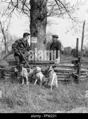 1920ER JAHRE SENIOR MANN IM GESPRÄCH MIT JÜNGEREN MANN JÄGER MIT SCHUSS PISTOLE UND DREI HUNDE JAGD GEBUCHTEN PLAKAT AM BAUM HAUSFRIEDENSBRUCH ZAUN Stockfoto