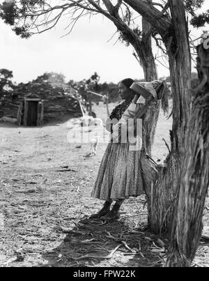 1930ER JAHREN INDIANISCHE NAVAJO FRAU HOLDING BABY PAPOOSE UNTER BÄUMEN MIT HOGAN IM HINTERGRUND Stockfoto