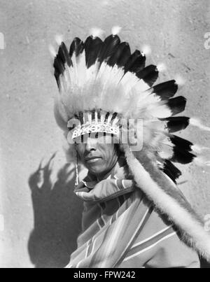 1930ER JAHREN INDIANISCHE MANN VOLL MIT GEFIEDERTEN KOPFSCHMUCK KRIEG MOTORHAUBE BLICK AUF KAMERA SAN ILDEFONSO PUEBLO NEW MEXICO USA Stockfoto