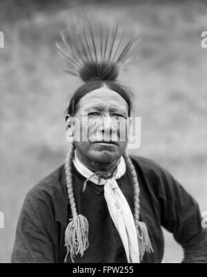 1920ER JAHRE PORTRAIT SCHÖNE INDIANISCHE INDISCHER MANN KOOTENAY STAMM TRÄGT FEDERN UND ZÖPFE BLICK IN DIE KAMERA Stockfoto