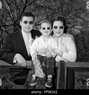 1950ER JAHRE FAMILIE VATER MUTTER TOCHTER AM ZAUN MIT DUNKLEN SONNENBRILLE BLICK IN DIE KAMERA Stockfoto