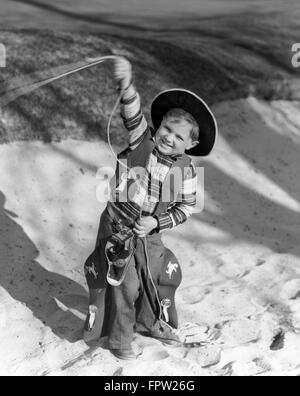 1930ER JAHREN LÄCHELNDE JUNGE IM COWBOY OUTFIT SCHWINGEN LASSO BLICK IN DIE KAMERA Stockfoto