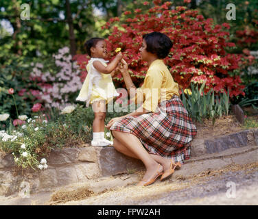 1980ER JAHREN LÄCHELND AFROAMERIKANISCHE FRAU UND KLEINE MÄDCHEN IM FREIEN IN DER NÄHE VON FRÜHLING BLUMEN TOCHTER GEBEN GELBE BLUME, MUTTER Stockfoto