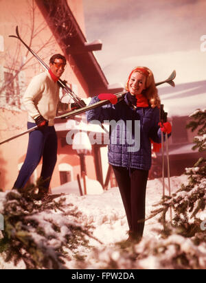 1970ER JAHREN PAAR MANN FRAU MIT SKIERN SKISTÖCKE SKI CHALET LODGE SCHNEE KIEFER BÄUME KLEIDUNG MODE LIFESTYLE Stockfoto