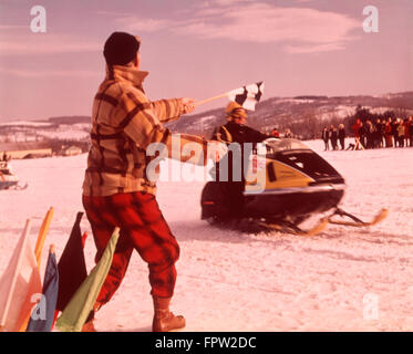 1960S 1970S SCHNEEMOBIL RENNEN MANN BEAMTER IN GELB ROT KARIERTEN WOLLE SCHNEE ANZUG WEHENDE ZIELFLAGGE KARIERTEN GEWINNER Stockfoto