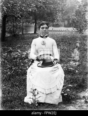 1890S 1900S PORTRAIT FRAU GESTREIFTEN KLEID SITZT IM STUHL IM GARTEN HOLDING IN RUNDE HUT Stockfoto