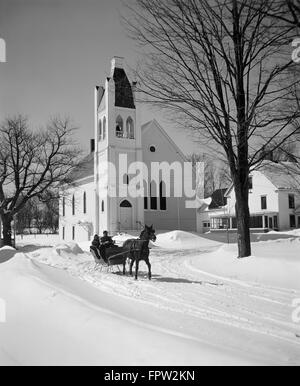 1960ER JAHREN PAAR TREIBENDE EINSPÄNNIGEN PFERDESCHLITTEN VOR LANDKIRCHE Stockfoto