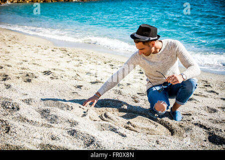 Junger Mann im schwarzen Hut sitzt am Strand und Sand berührt, während wegschauen Stockfoto