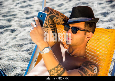 Schönen jungen Mann am Strand während der Einnahme von Selfie Foto mit Handy auf Liegestuhl sitzen. Seelandschaft mit Hügeln im Hintergrund Stockfoto