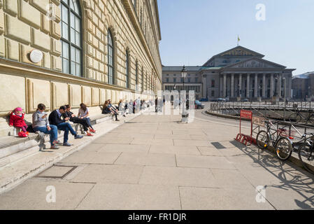 Leute sitzen an der königlichen Königsbau Palast und Theater in München genießen die warme Sonne im Frühling, Bayern, Deutschland Stockfoto