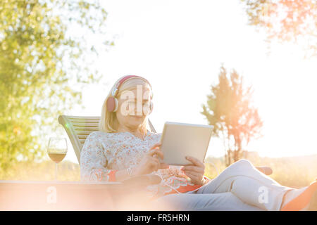 Ältere Frau mit Kopfhörer und Wein mit digital-Tablette auf Lounge-Sessel in sonnigen Hinterhof Stockfoto