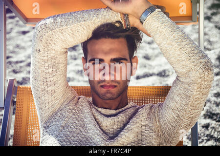 Schönen jungen Mann am Strand sitzen auf Liegestuhl ausruhen und den Urlaub genießen. Tragen von leichten weißen Pullover Stockfoto