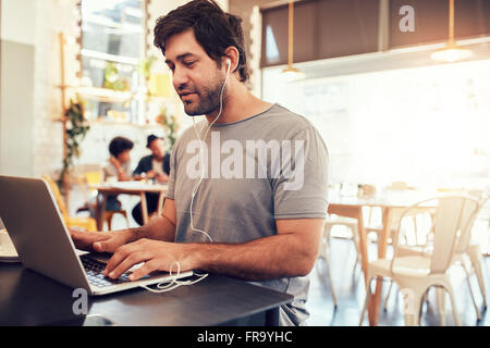 Porträt von hübscher junger Mann mit Kopfhörer am Laptop sitzen in einem Café arbeiten. Junger Mann an einem Café Surfen inte Stockfoto