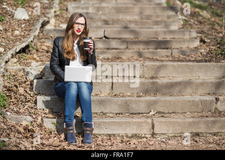 Junge hübsche Frau mit Laptop mit Kaffeepause auf der Treppe sitzt. Kaukasische Mädchen arbeiten mit Notebook in der Natur. Stockfoto