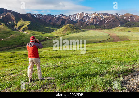 Ansicht eines älteren Mannes in einer orangen Jacke, der ein Bild von Alaska Range, Grant Creek und der Ebene unter dem Highway Pass mit üppiger grüner Tundra und... Stockfoto