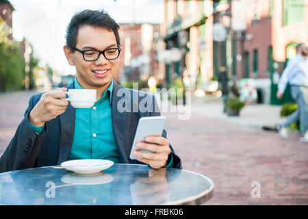 Junge asiatische Mann in casual Geschäftskleidung lächelnd in entspannenden outdoor Cafe Tasse Kaffee zu trinken, beim Verwenden von mobile Stockfoto