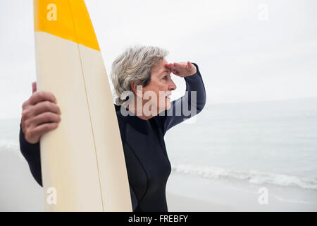 Ältere Frau mit Surfbrett Abschirmung Augen am Strand Stockfoto