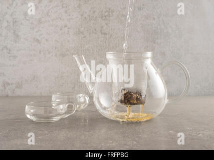 Heißes Wasser im Glas Teekanne gießen und Sichten Tee Blätter. Zwei Tassen aus Glas befinden sich auf der Seite auf einem Stein Küchentisch Stockfoto