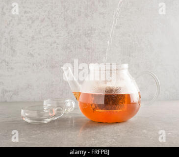 Heißes Wasser im Glas Teekanne gießen und Sichten Tee Blätter. Zwei Tassen aus Glas befinden sich auf der Seite auf einem Stein Küchentisch Stockfoto