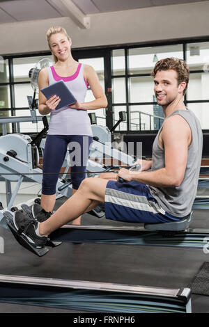 Porträt von Trainer schreiben auf einem Klemmbrett während Mann Training auf einem Rudergerät Stockfoto