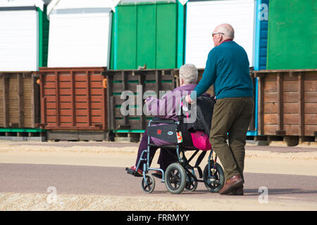 Mann, die Frau im Rollstuhl Promenade vorbei Strandhütten bei mittleren Chine, Bournemouth im März schieben Stockfoto