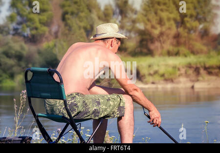 An den Ufern des Flusses auf einem Klappstuhl sitzt ein Angler mit Angelrute und Fisch zu fangen. Stockfoto