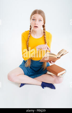 Erstaunt Studentin sitzen auf dem Boden mit Buch und Blick in die Kamera auf einem weißen Hintergrund isoliert Stockfoto