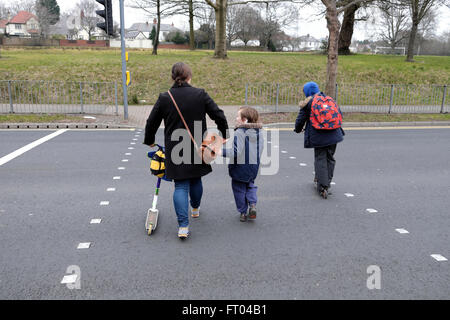 Mutter Wandern mit Kindern und Roller zur Schule Überqueren einer Straße an einem fußgängerüberweg, bevor es in Wales UK KATHY DEWITT zu arbeiten Stockfoto