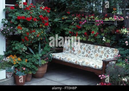 Geranien in Töpfen, um und über eine Holzbank mit einem gemusterten Kissen auf einem 70er Jahre Terrasse Stockfoto