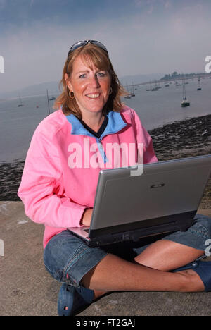 Eine Frau in einer rosa Fleece gekreuzt Sitzen im Schneidersitz auf den Strand in Beaumaris, Anglesey, Wales, Großbritannien, mit Ihrem Laptop. Stockfoto