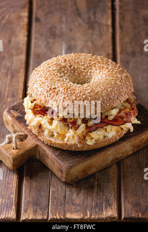 Bagel mit Speck, Rührei und gebratenen Zwiebeln auf kleine hölzerne hacken an Bord über hölzerne Hintergrund. Stockfoto