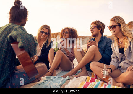 Gruppe von jungen Leuten anhören Freund Gitarre im Freien zu spielen. Heterogene Gruppe von Freunden abhängen am Strand. Junge Männer und