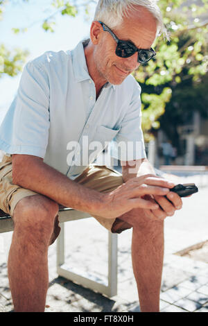Porträt von senior glücklich mit Handy beim Sitzen auf einer Bank in der Stadt hautnah Stockfoto